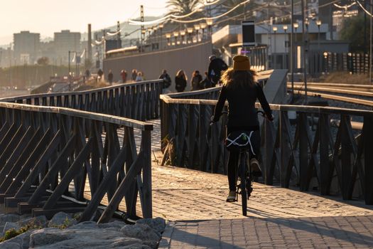 Young girl with curly hair fluttering in the wind is riding a bicycle on wooden bridge at sunset on the promenade road of Maresme, Catalonia, Spain.