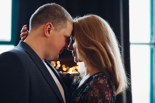 Elegant couple touching each other by heads, face to face, with closed eyes. Boyfriend wearing in dark blue jacket and blonde girlfriend in flowering chiffon dress on wooden background with lights.