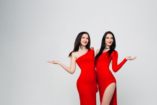 Studio portrait of beautiful happy twins with red lips and long brunette hair embracing in red clothes and smiling at camera over white isolated background.