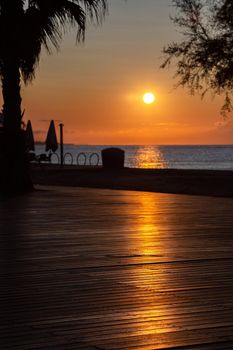 Orange sun rising above the sea. View from promenade with the sea, beach and cafe. The solar path is reflected from pedestrian zone made of wood.