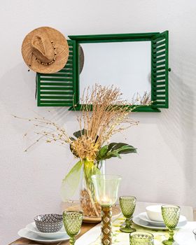 Fragment of nicely decorated and served living, lunch room with the mirror and straw hat. A table served with plates, glasses and decorative flowers in vase. Interior design. Vertical.