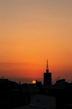 Orange summer sunrise or sunset, roof top view of an old church tower above the city. Warm red, yellow and orange sky color tones. Old town of Badalona, Spain