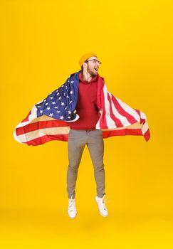 Happy, funny young man in red hoodie and USA flag jumping hight isolated on yellow background. freedom is in your life. Cheerful young man with American flag.