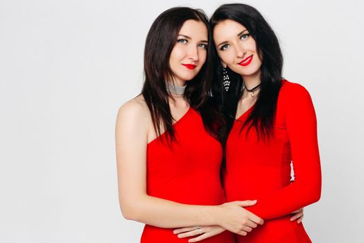 Two sexy and beautiful sisters twins in red dresses posing, looking at camera holding hands up. Pretty dancing ladies with long hair. Fashionable and stylish girls. Shopping, fashion, party.