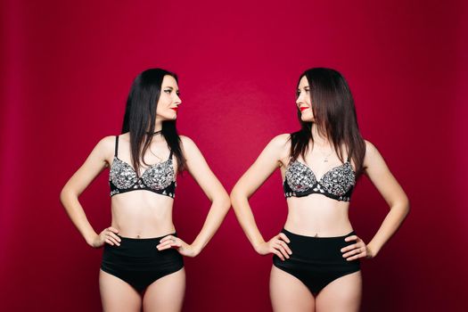 Studio portrait of sexy beautiful twin girls in striptease costumes smiling face to face with hands on hips. Isolated on red background.