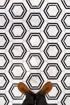 Top view of men's legs in black jeans wear brown leather shoes, standing on black and white hexagon floor tiles.