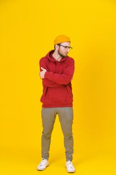 Pensive young man in casual wear in glasses posing looking sideways on yellow background with one arms folded. Stylish bearded smart hipster man casual look. Thoughtful guy on yellow background.