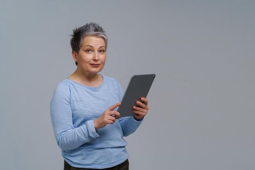 Marvelous mature woman 50s grey haired with digital tablet working or checking on social media. Pretty woman in blue blouse isolated on white. Older people and technologies. Toned image.
