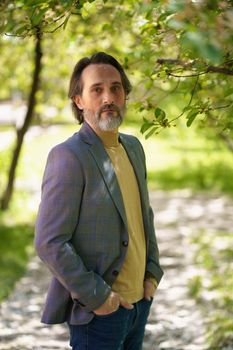 Mature handsome man with grey beard looking thoughtful at the camera wearing casual jacket and jeans holding hands in pockets.