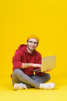 Happy young hipster man in red hoodie sitting on the floor holding laptop studying or working. Smart freelancer caucasian man with laptop isolated on yellow background.