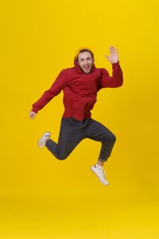 funny portrait of a young man in joy jumping high wearing casual red hoodie and jeans isolated on yellow background. Humorous hipster guy in jump.