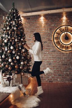 Side view full length of slim young girl with long dark hair in white sweater, black leggings and white socks decorating Christmas tree at home . Clock on brick wall shows almost midnight.