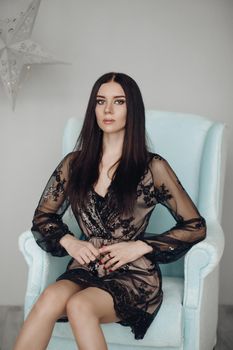 Full length studio portrait of attractive young brunette in evening laced gown and high heels sitting in frozen blue arm-chair. Room is decorated with paper black and white stars in background.
