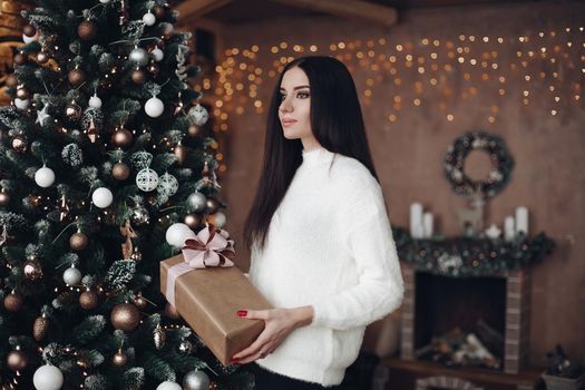 Portrait of attractive young woman with long black hair and evening make up wearing cozy white sweater holding wrapped Christmas or New Year present. She is in decorated room with Christmas tree and fireplace with illuminated garland above.