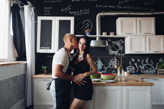 View from side of beautiful pregnant woman standing near caring husband at kitchen. Beloved man cooking dinner for adorable wife at home. Future mother and father expecting baby. Concept of love.