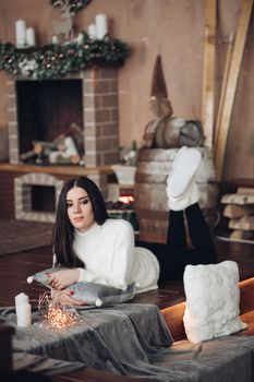 Portrait of stunning attractive young woman with long dark hair and bright evening make up in white sweater sitting on the floor with christmas gifts. She is holding white toy dwarf. Looking at camera.