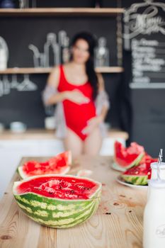 Focus on cut juicy watermelon on wooden table. Unfocused blurred background with unrecognizable pregnant brunette woman in red embracing her belly.
