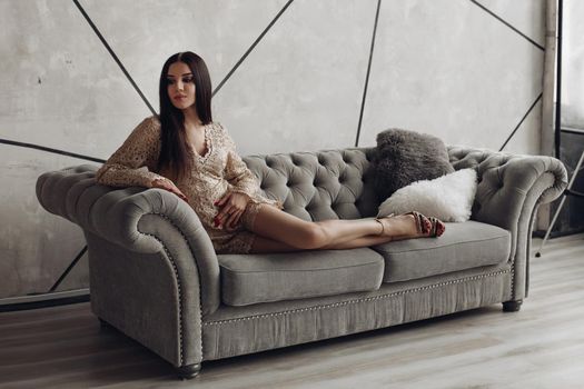 Full length of stunning brunette young model in beige laced romper and high heels posing on luxurious grey sofa with furry pillows.