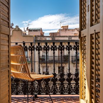Balcony with a wrought iron railing, wooden sashes and wicker chair. Sunny day with blue sky. View to a building in Barcelona, Spain. Vacation and travel.