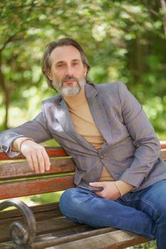 Mature handsome freelancer man with grey beard, looking thoughtful at the camera sitting on the bench at the park wearing casual jacket and jeans holding smartphone in hand.