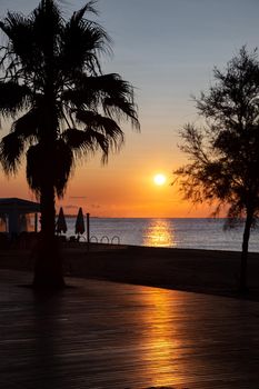 Orange sun rising above the sea. View from promenade with the sea, beach and cafe. The solar path is reflected from pedestrian zone made of wood.