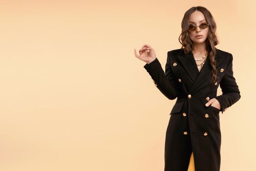 Full length fashionable portrait of gorgeous young brunette woman in trendy sunglasses, black double-breasted coat and black high heels. Wavy hair, looking at camera. Isolate on plain background.