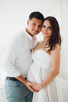 Happy future mother and father hugging each other, looking at camera and smiling. Attractive pregnant woman in white dress awaiting little baby. Concept of pregnancy and happiness.