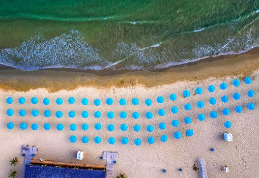 Aerial top view of a beautiful small beach with blue umbrellas, and sea waves.