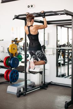 Back view of young sporty and strong girl with athletic body doing exercises on bar in gym. Brunette woman with short hair, doing workout tightening on bar. Concept of sport.
