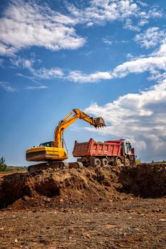 Yellow excavator and empty dump truck working at the construction site. Vertical view