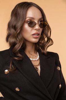 Brunette model with curly hair standing with slightly open mouth. Surprised young girl in sunglasses and black jacket posing on light studio background. Gorgeous lady wearing jewelries on neck.
