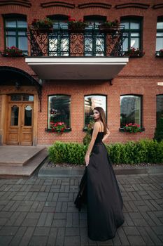 Portrait of a luxurious woman in a lush long black dress. The model is represented by a view from behind with a pensive look to the side. Against the background of a beautiful building.