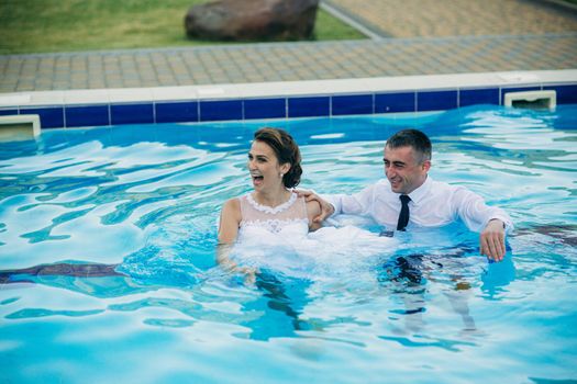 Young couple jumping in the swimming pool in a wedding suit and wedding dress. Sunny day.