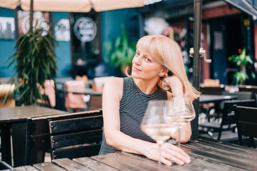 Mature adult woman sitting in bar outdoors with wine glasses and blurry restaurant background scene, drinking white wine and eating cheese. Summer sunny day on patio. People lifestyle