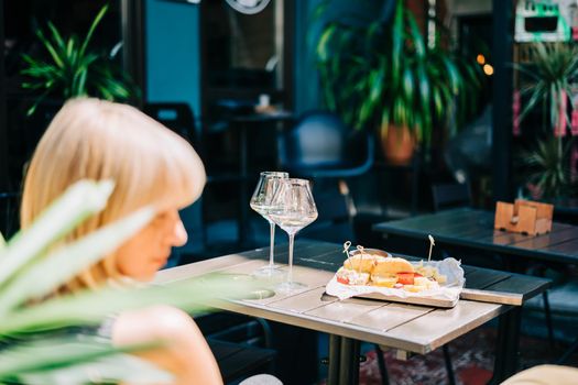 Young woman sitting in cafe outdoors with glasses and blurry restaurant background scene, drinking white wine and eating cheese. Summer sunny day on patio outdoors.