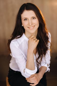 Portrait of a happy young brunette woman in a white shirt sitting on a chair.