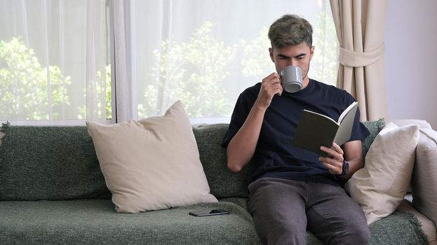 Millennial man drinking hot coffee and reading book on sofa at home.