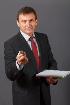 Confident and friendly elegant handsome mature businessman standing in front of a gray background holding a marker and a notebook