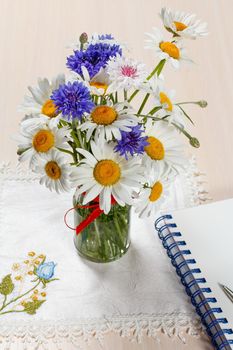 Daisy chamomile and cornflowers in vase on a wooden table with a napkin and a notebook