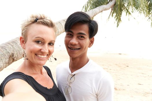 Adult caucasian woman taking a selfie with her asian male couple in a tropical beach