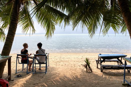 Multicultural couple sitting on chairs in a tropical beach looking to the horizon while talking