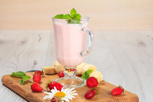 Glass of delicious milk shake with mint and fresh strawberries, banana, chamomile on a wood cutting board