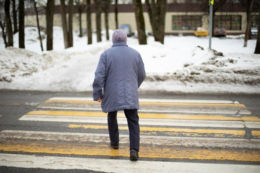 Woman crosses road on zebra. Pensioner in Russia walks down street. Man walks on pedestrian crossing. Shooting from back.