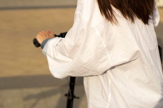 A girl in a loose white jacket rides on a black scooter, close-up, side view. A shot with long hair. Photo in motion. Walks around the city. Outside shooting