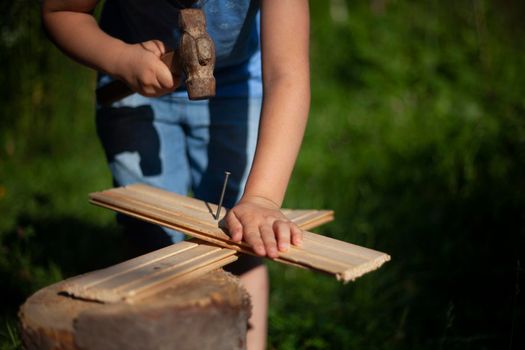 Child hardens nail into plank. Girl learns to beat with hammer. Preschooler is engaged in manual labor. Learning without computer. Work in fresh air. Playing with instruments.