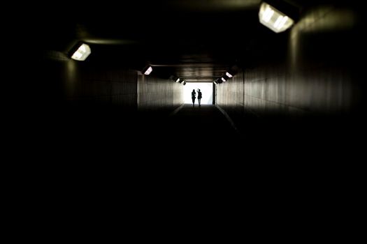 Two silhouettes at end of tunnel. Light in distance. Figures of small women in contrasting light. Lamps in long underpass.