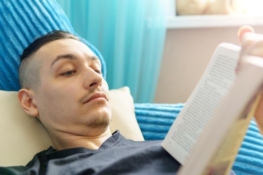Man thoughtfully reading book comfortably lying on sofa. Selective focus