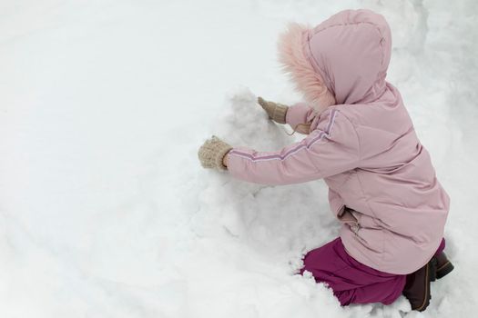 Child sculpts snowman. Girl plays in snow. Baby in winter on street. Preschooler in warm clothes plays alone.