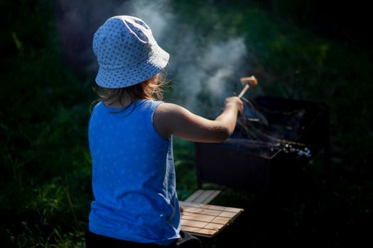 Child at picnic. Girl bakes bread on stick. Summer outdoor recreation. Child near fire. Child in panama. Girl holds stick of bread over fire.