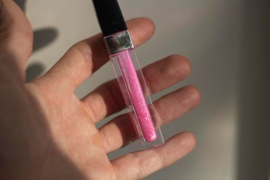 A bottle with a pink lip gloss lies in the palm of a person's hand, illuminated by sunlight. Makeup tool. Indoor shooting